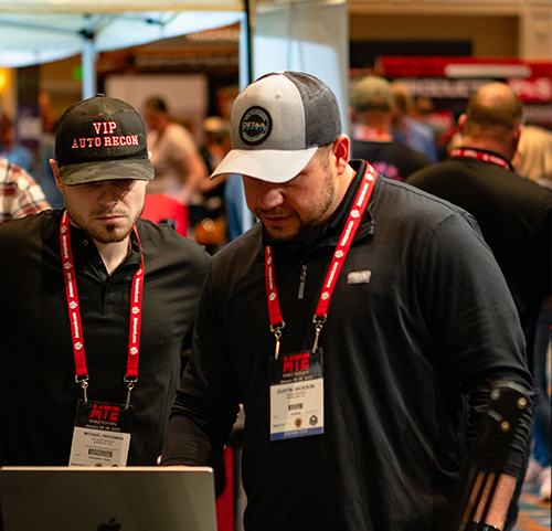 Two men wearing caps and lanyards look at a laptop screen at an indoor event.